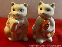 Front view of the pair of cat-shaped porcelain teapots on red surface, showing detailed faces and painted flowers.