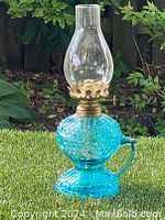 Full view of vintage blue glass oil lamp on grass background showing entire lamp with clear chimney and gold-tone burner.