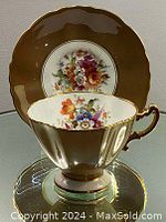 Teacup displayed inside saucer showing hand painted floral design, gold trim on handle and scalloped rim in a brown base color.