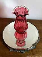 Top view showing cranberry glass vase reflected in round beveled mirror with ornate rococo floral metal border.