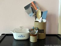 Photo showing complete kitchen lot with stoneware containers, souvenir spoons, casserole dish, and utensils arranged on black surface.