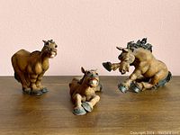 Three comical brown resin horse figurines on wooden surface against pale pink wall. Left horse standing with wide grin, middle horse reclining with closed legs, right horse bucking on raised legs.