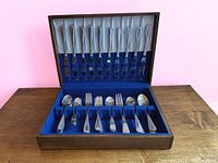 Open wooden case displaying 12 knives in top section with blue felt backing and lower compartment holding forks and spoons organized into slots.