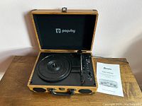 Front view of Popsky suitcase turntable opened, showing turntable platter, tonearm, and user manual beside it on table.