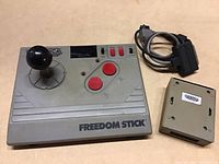 Top view of the Cameroca Freedom Stick joystick showing joystick, buttons labeled A and B, switch, and cable ending in 9-pin connector, alongside an adapter unit.