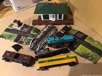 Various vintage HO scale train cars and accessories arranged on a surface including station building, track sections with scenery base, bridge, and multiple train cars