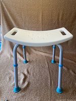 Front angled view of the white plastic adjustable shower safety seat showing the shape of the seat with drainage holes and cutout handles, as well as the adjustable aluminum legs with blue adjustment rings and rubber feet.