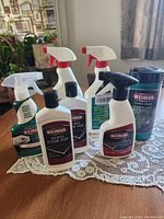 Photo of full set of seven cleaning products including 2 Woodpecker floor cleaners, 2 granite/stone/marble cleaners, and 3 glass cooktop cleaners on lace tablecloth.