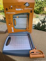 Paper trimmer with steel blade set on an outdoor table with original orange Fiskars box behind it showing product image and specifications.