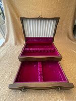 Open flatware chest showing white pleated fabric on lid interior, red velvet flatware holder slots and lower velvet-lined drawer with divider.
