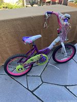 Side view of girls' bike showing purple frame, pink tire rims, green chain guard, adjustable white seat with floral pattern, and handlebar streamers.