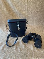 Binoculars and carrying case displayed on a textured fabric surface, showing the capped lenses with the Kurt Müller logo and attached leather strap.
