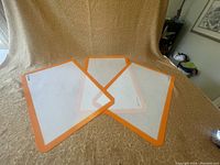 Three rectangular Silpat silicone baking mats with orange borders laid out on textured fabric.