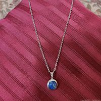 Full view of silver chain necklace displayed on red striped fabric, showing pendant with round blue stone.