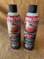 Front view of two black and red cans of FastWax FW1 Cleaning Wax spray on a textured surface