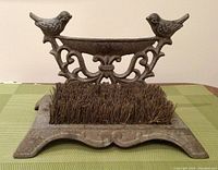 Front view showing the iron boot scraper with two birds perched on a decorative birdbath structure and bristles for scraping boots.
