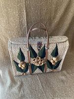 Front view of vintage rattan tote bag showing woven texture, leather handles, floral leaf, and flower embellishments on flap with metal clasp closure.