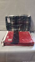 Lot consists of two folded Horn Brothers blankets in plastic packaging. Red blanket visible on top foreground with black central stripe and white edging; green plaid blanket with blue and white accents folded behind.