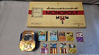 Monopoly game box and metal tin with Pokémon cards laid out, showing several card front faces.