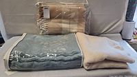 Four blankets stacked on a surface: off-white folded, light blue blanket, and two light brown blankets packed in plastic with tassels visible