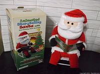 Animated Storytelling Santa plush figurine sitting in a white rocking chair holding an open storybook, next to original box showing front design with fireplace and Christmas tree illustration.