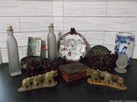 Wide view of lot showing all items including figurines, plate, vase, wooden box, and glass bottles on black surface against wood patterned background