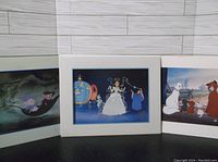 Three Disney lithographs standing side by side on dark floor with white wall background: The Rescuers, Cinderella, and The Aristocats