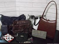 Photo showing all 8 assorted handbags and wallets arranged together: includes dark brown leather shoulder bag, burgundy textured leather bag with top handle, cream and black studded tote, navy blue shoulder bag, black fabric crossbody bag, gray wallet, small floral zippered wallet, small dark blue wallet.