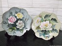Front view of two floral hand painted porcelain plates with scalloped edges and gold outlines. One has the image of pink, yellow, and white roses on a blue background, the other a white and cream flower on green background.