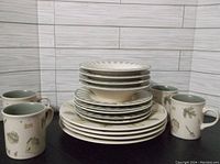 Stacked Pfaltzgraff Naturewood dinnerware including plates, bowls, and mugs arranged on a black surface with light wood panel background.