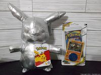 Silver Pikachu plush toy with Pokemon tag, placed next to a sealed Pokemon TCG Cosmic Eclipse booster pack with promo card and coin in packaging.
