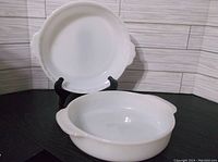 Two white milk glass pie plates displayed. One Pyrex #228 plate shown standing on black stand, one Fire King round pie plate in front.