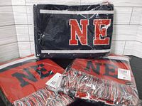 Photo shows three packed New England scarfs, two with red backgrounds and one navy blue, featuring large 'NE' letters and fringed ends on two scarfs.