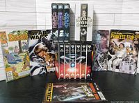 Photo showing Star Wars and Star Trek VHS tapes arranged upright with Star Wars comics laid flat in front, plus a set of Star Wars Trilogy VHS tapes displayed horizontally.
