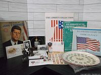 Photo showing an assortment of JFK memorabilia including 'The Story of John F. Kennedy' book, Liberty Collection documents, a sealed decorative plate, a White House ornament, and several black and white photos and figurines.