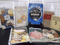 Eight vintage softcover cookbooks arranged on a surface, displaying cover titles and artwork focused on cakes and desserts.