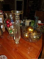 Photo showing tall crystal candlestick, glass vase filled with holiday ornaments, and crystal bowl with gold balls on wooden table