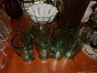 Eight dark green Coca-Cola glasses on a wooden table, showing varied shapes including tall cylindrical and mug styles.