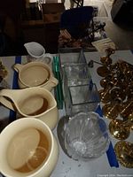 Photo showing the cream ceramic pitchers and vases alongside square glass vases and brass items on a table.