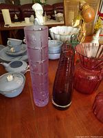 Two hand blown heavy crystal vases displayed on a wooden table alongside various ceramics and decorative objects, clearly showing the purple cylindrical vase with rectangular cuts and a red vase with grid patterns.