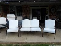 Four-piece outdoor seating set with white cushions on chairs and love seat placed on concrete patio in front of brick wall and windows.