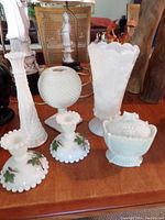 Various milk glass items including vases, bowls, and candle holders with hand painted details arranged on a wooden surface.
