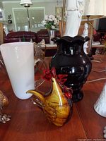 Overview of the hand blown rooster figurine, black vase, white vase, and clear glass pitcher displayed on a wooden table.