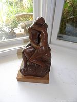 Photo showing front and side of The Kiss sculpture on wooden base near window with outdoor garden view.