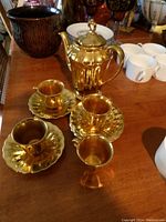 Gold-tone tea pot, four cups, and three saucers displayed on table with other items in background.