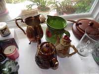 Overview of all pitchers and teapots together on window sill including sterling plated, ceramic, and glass items