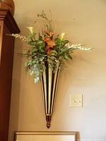 Two large wall sconces mounted on a wall, each holding floral arrangements with mixed flowers and greenery. The sconces have a black and gold striped vase shape and decorative base.