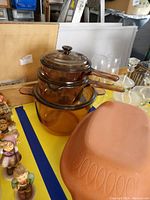 Two small amber glass saucepans with lids stacked on a larger amber glass pot, with a terracotta lidded clay pot and various items around.