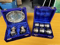Two open blue velvet boxes containing antique silverware sets placed on a table; left box contains salt and pepper shakers with a plate in the lid, right box contains six napkin rings.