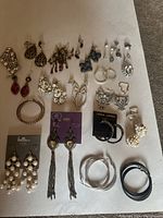 Wide shot of 18+ pairs of earrings and some single studs laid out on white surface showing different styles including pearls, rhinestones, hoops, and colorful dangles.